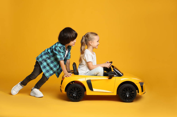 Cute boy pushing children's electric toy car with little girl on yellow background - Фото, зображення