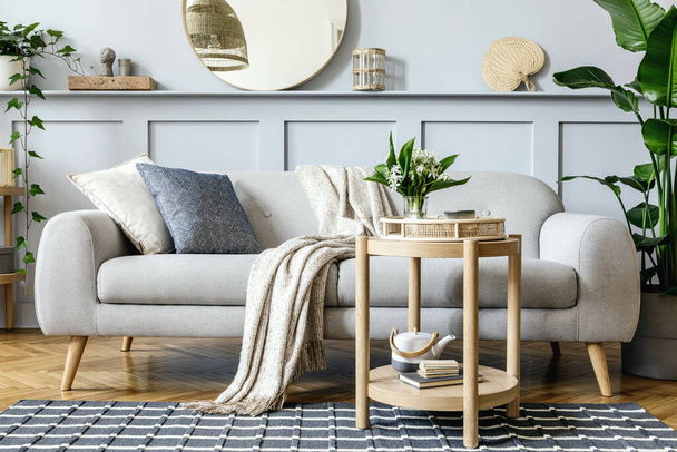 Scandinavian living room interior with design grey sofa, wooden coffee table, tropical plants, shelf, mirror, furniture, plaid pillow, teapot, book and elegant personal accessories in home decor. - Φωτογραφία, εικόνα