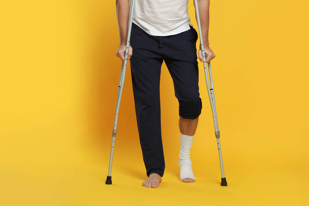 Man with injured leg using crutches on yellow background, closeup - 写真・画像
