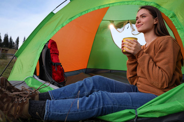 Young woman with cup of drink in camping tent outdoors - Фото, изображение