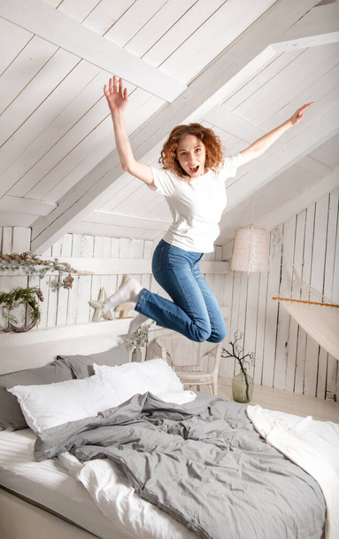 European young  fun woman with red hair in jeans jumps on a bed smile,  in a bright room with a Christmas interior, - Foto, Imagem
