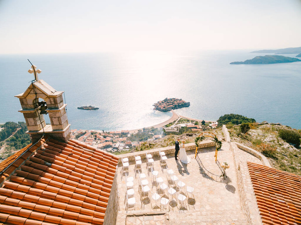 Braut und Bräutigam stehen auf der Aussichtsplattform vor der Kirche Sveti Sava mit Blick auf die Insel Sveti Stefan. Montenegro. Blick von oben - Foto, Bild