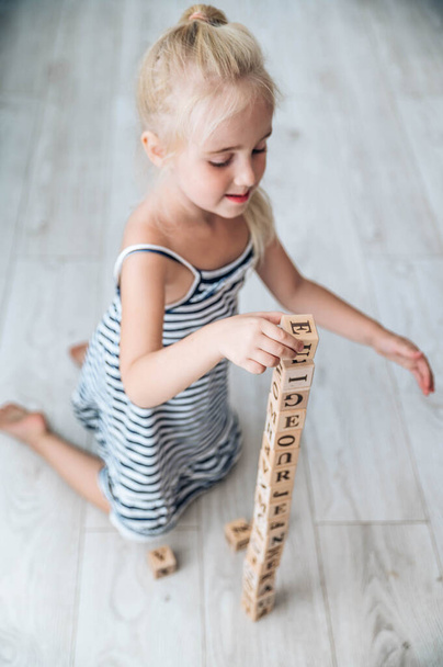 Маленька мила блондинка грає і навчається з кубиками абс в приміщенні. Маленька дівчинка грає з дерев'яними блоками на підлозі
 - Фото, зображення