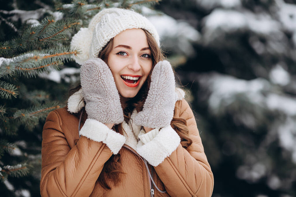 Удивлённая девушка, улыбающаяся в камеру в зимнем лесу в тёплой коричневой куртке - Фото, изображение