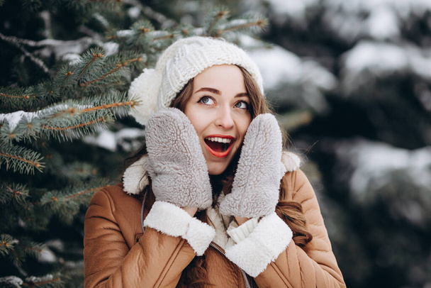 Chica sorprendida sonriendo a la cámara en el bosque de invierno con una chaqueta marrón cálido y sombrero blanco - Foto, imagen