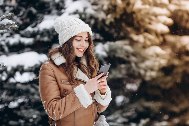 Kış parkında telefonuna gülümseyen mutlu bir kızın portresi. Üzerinde kalın bir ceket ve beyaz bir şapka vardı. - Fotoğraf, Görsel