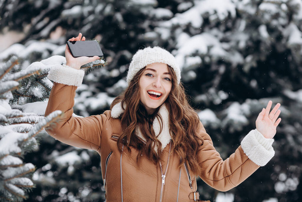 Portrait of a happy girl smiling to camera and holding her phone in a winter park wearing a warm jacket and white hat - Photo, Image