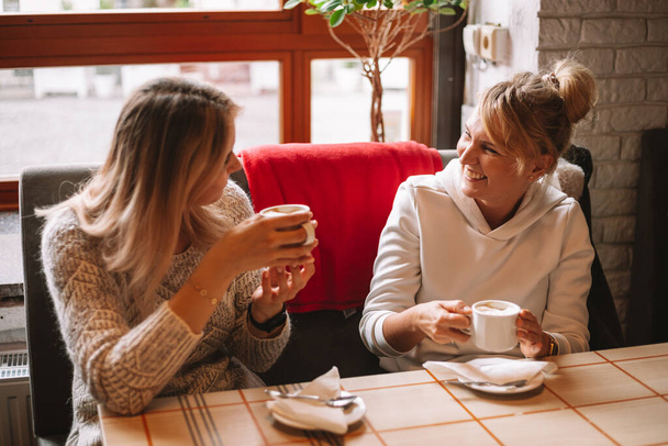 Photo de deux jeunes femmes blanches heureuses avec des tasses à café au café, avoir une conversation, rire, moments drôles, mode de vie. Belle conversation entre amies - Photo, image