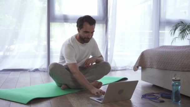 家でフィットネスマットで瞑想しながら、ラップトップを使用して男  - 映像、動画
