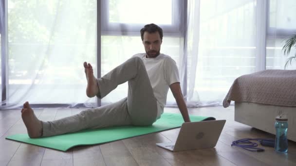 Mężczyzna ćwiczy na macie fitness w pobliżu laptopa w sypialni  - Materiał filmowy, wideo