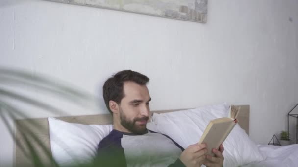 Ευτυχισμένος άνθρωπος διαβάζει το βιβλίο, ενώ βρίσκεται στο κρεβάτι στο σπίτι  - Πλάνα, βίντεο
