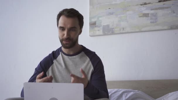 Mosolygós férfi gesztikulál, miközben a videó hívás laptop a hálószobában  - Felvétel, videó