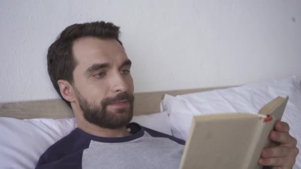 Uomo barbuto sorridente durante la lettura del libro sul letto  - Filmati, video