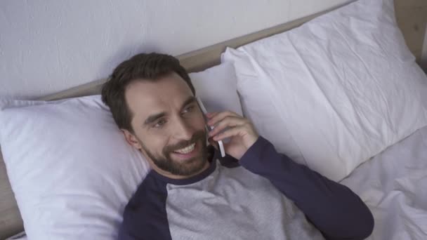 Vista superior del hombre alegre hablando en el teléfono inteligente en la cama  - Metraje, vídeo