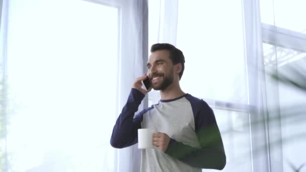 Χαμογελώντας άνθρωπος με κύπελλο μιλάμε στο smartphone στο σπίτι  - Πλάνα, βίντεο