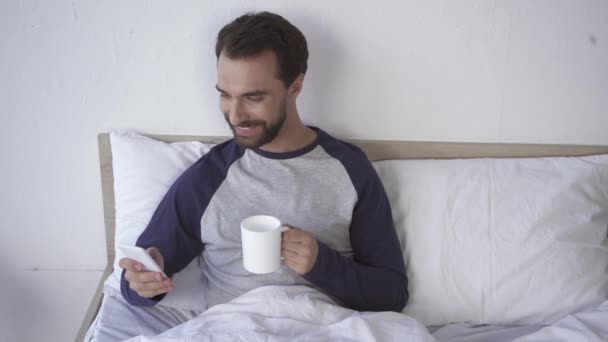 Χαρούμενος άνθρωπος χρησιμοποιώντας smartphone και κρατώντας κύπελλο στο κρεβάτι  - Πλάνα, βίντεο