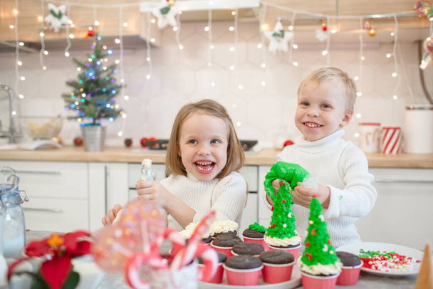 Little children prepare a Christmas treat in the white kitchen. The boy and the girl are dressed in white sweaters. Magic moment - Foto, afbeelding