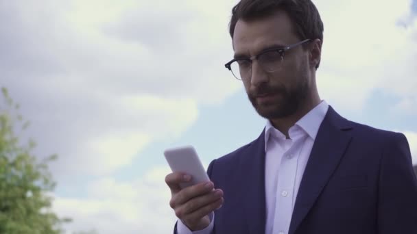 Empresario en gafas de vista sonriendo y usando smartphone al aire libre  - Metraje, vídeo