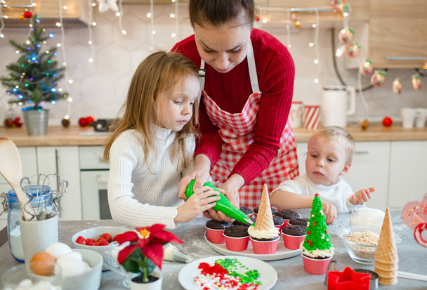Cute family prepare a Christmas treat in the white kitchen. The boy and the girl are dressed in white and their mother in red sweaters. Magic moment - Foto, Imagem