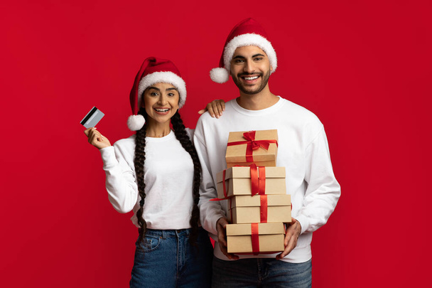 Χριστουγεννιάτικα ψώνια. Ενθουσιασμένο αραβικό ζευγάρι με πιστωτική κάρτα και στοίβα από δώρα - Φωτογραφία, εικόνα