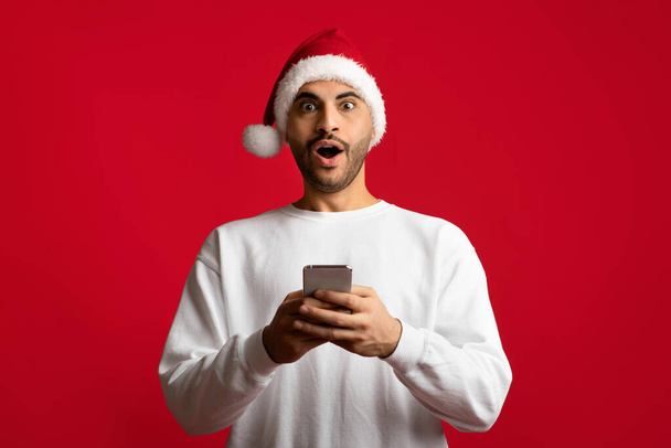 Erstaunliches Angebot. Porträt eines überraschten arabischen Mannes mit Weihnachtsmütze und Smartphone - Foto, Bild