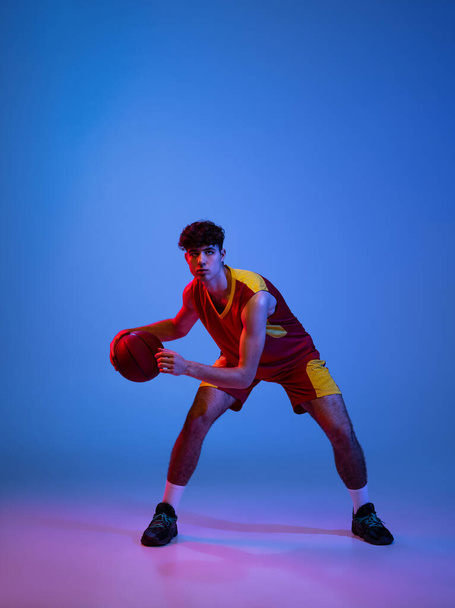 Studio shot of young man, professional basketball player playing basketball isolated on blue background in neon light. - Φωτογραφία, εικόνα