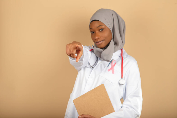 Foto di medico musulmano donna che punta con indice verso la fotocamera avvolta in velo grigio e cartella con volto triste. - Foto, immagini