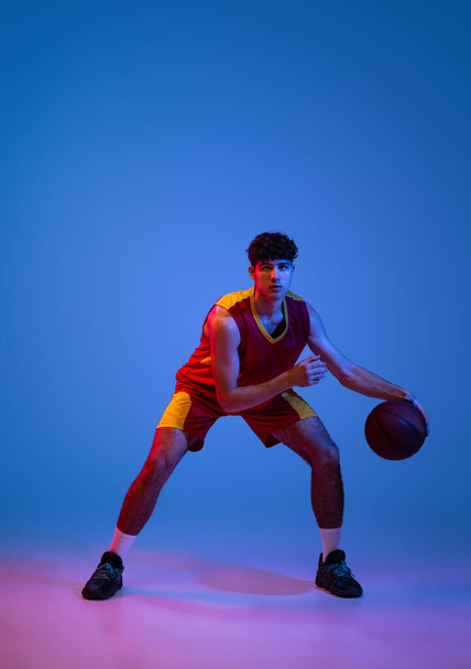 Studio shot of young man, professional basketball player playing basketball isolated on blue background in neon light. - Фото, зображення