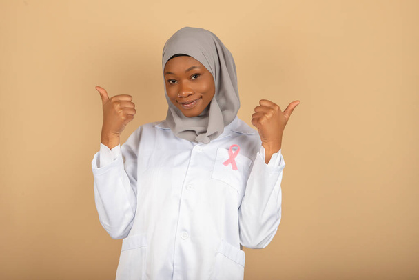 Muzułmańska lekarz w chustce i białym płaszczu nosi różową wstążkę świadomości raka piersi, podnosi rękę z kciukiem w górze - Zdjęcie, obraz