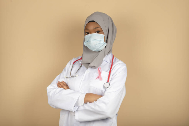 Portret afroamerykańskiej lekarki w masce chirurgicznej, szaliku i białym płaszczu, pozującej z skrzyżowanymi rękami. - Zdjęcie, obraz