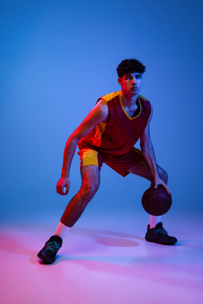 Studio shot of young man, professional basketball player playing basketball isolated on blue background in neon light. - Photo, Image
