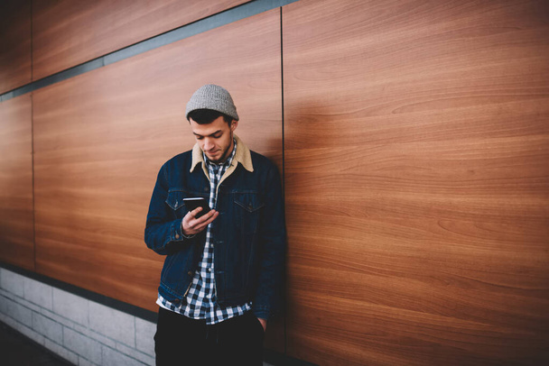 Pensive male in warm clothes standing near wooden wall with hand in pocket while typing message on mobile phone during break - Foto, Imagen