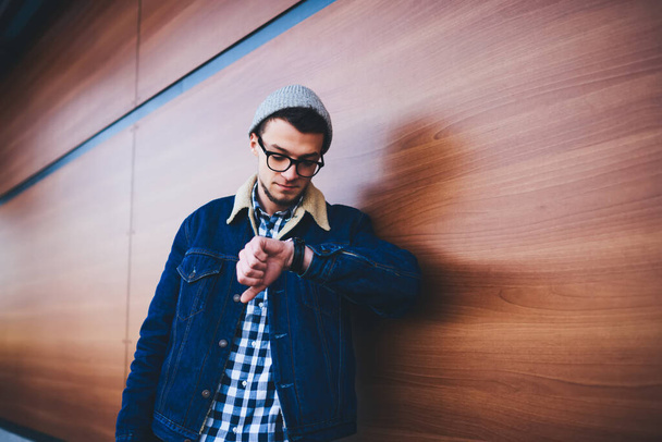 Mladý vousatý muž v neformálním oblečení a brýlích opírající se o dřevěnou zeď na ulici a kontrolující čas na hodinkách při čekání na partnera - Fotografie, Obrázek