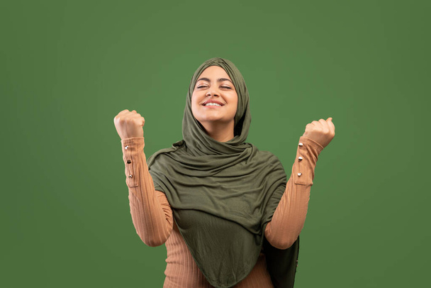 Євфорична арабська леді в хіджабі святкування успіху з піднятими руками і згорнутими кулаками, темно-зелений фон
 - Фото, зображення