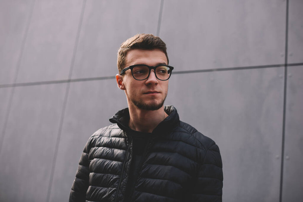 Low angle of thoughtful young man in dark jacket and eyeglasses standing near modern building on street and looking away - Fotografie, Obrázek