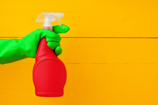 Hand in glove holding household detergent spray against yellow wooden background - Zdjęcie, obraz