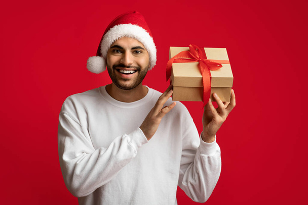 Счастливого Рождества. Возбужденный араб в Санта-Шляпе, держащий упакованный ящик с подарками - Фото, изображение