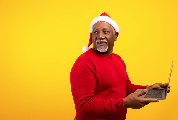 Senior μαύρο άνδρα χρησιμοποιώντας φορητό υπολογιστή για ψώνια Χριστούγεννα, κοιτάζοντας πίσω από τον ώμο του σε κενό χώρο για τις διακοπές διαφήμιση - Φωτογραφία, εικόνα
