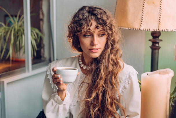 Portret van een jonge vrouw met een vintage blouse die in het café thee of koffie drinkt. Selectieve focus. - Foto, afbeelding