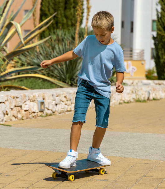 Preschooler kid boy in blue t-shirt learning to ride a skateboard outdoors. Cute child doing sports and having fun in park. Active hobbies, healthy lifestyle concept. T-shirt mockup - Foto, Imagem