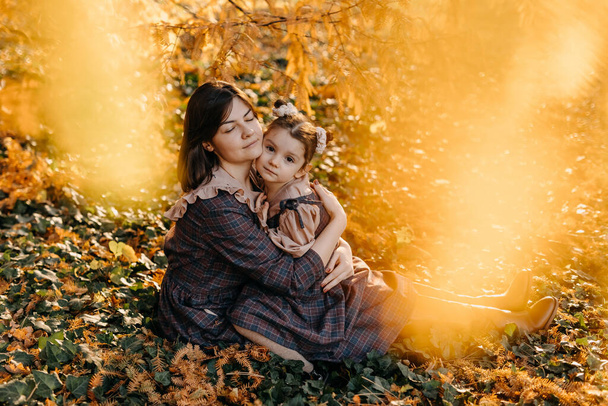 女性と少女、抱擁、公園で地面に座っている。秋の日に屋外で家族で過ごす母と娘. - 写真・画像