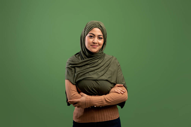 Muzułmańska koncepcja piękna. Portret młodej arabki w hidżabie pozującej ze złożonymi ramionami na ciemnozielonym tle - Zdjęcie, obraz