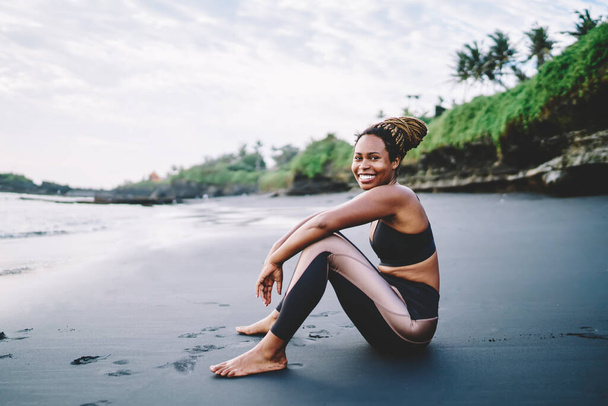 Happy African American resting at beach seashore and smiling at camera during time for morning workout on nature, portrait of cheerful female jogger in tracksuit enjoying body positive lifestyle - Foto, imagen