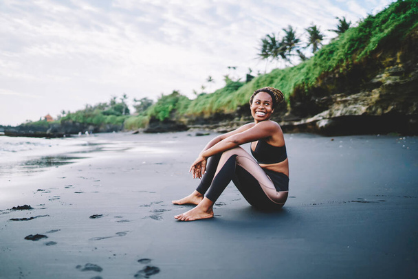 Feliz afroamericano descansando en la orilla del mar y riendo durante el tiempo para el entrenamiento matutino en la naturaleza, alegre jogger femenino en chándal disfrutando de un estilo de vida corporal positivo relajándose en la costa - Foto, imagen