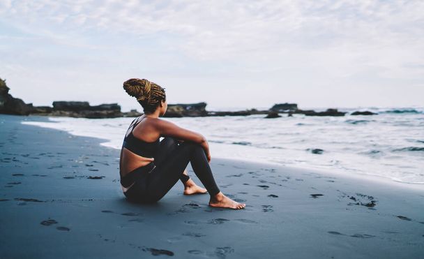 Dark skinned female runner with casual figure resting at coastline thinking about body positive lifestyle, African American woman in tracksuit spending morning time for training at seashore - Φωτογραφία, εικόνα