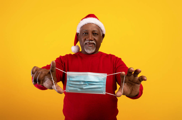 Szczęśliwy starszy czarny mężczyzna w kapeluszu Mikołaja trzymając maskę twarzy w wyciągniętych rękach, świętując Boże Narodzenie bez koronawirusu - Zdjęcie, obraz