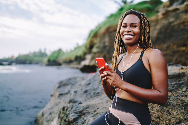 Happy African American runner with dreads enjoying audio songs via earphones connected to smartphone, funny dark skinned jogger in active clothes for training listening music from cellphone - Foto, afbeelding