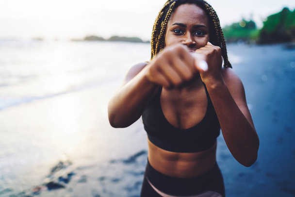 Half length portrait of African American sportive girl in active wear making boxing exercises to improve shape of hands during morning sport training at sea coastline, concept of endurance - 写真・画像