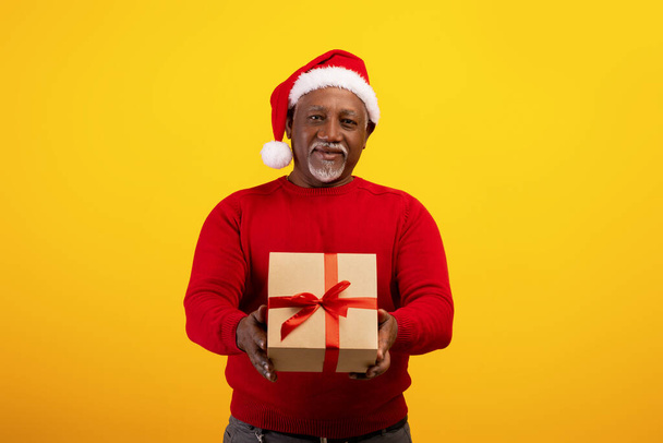 Portret wesołego starszego czarnego mężczyzny w kapeluszu Świętego Mikołaja trzymającego zapakowany świąteczny prezent na pomarańczowym tle studia - Zdjęcie, obraz