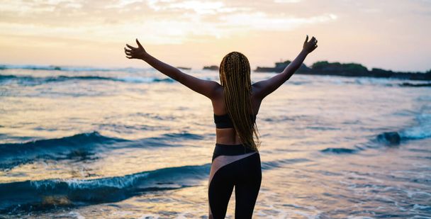 Back view of satisfied female runner in sportive tracksuit raising hands during workout break at seashore coastline, carefree woman with casual figure enjoying slimming goals and own triumph - Valokuva, kuva